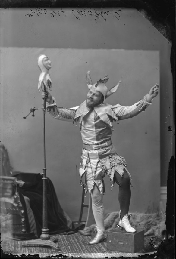 Photo noir et blanc d’un homme costumé en fou du Roi prenant la pose dans un studio de photographie. Il tient une marionnette sur pied dans sa main droite.