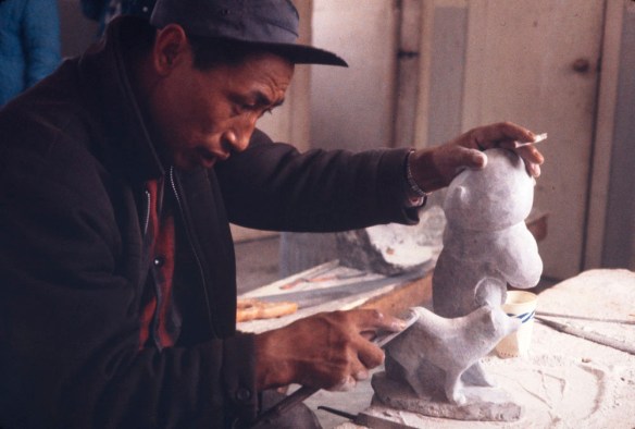 Photographie couleur d’un homme inuit portant une veste et une casquette foncées, en train de sculpter des statues blanches.