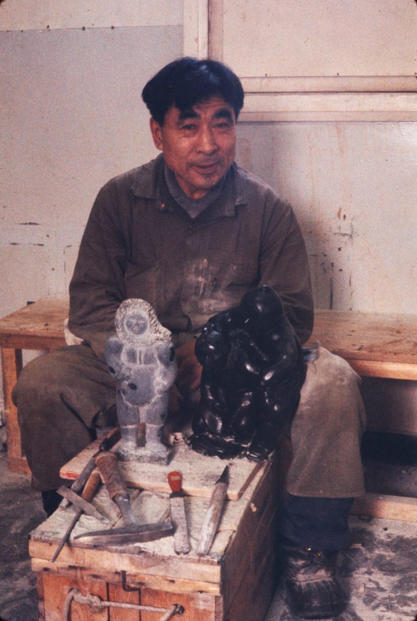 Photographie couleur montrant un homme inuit assis derrière une sculpture de pierre et des outils.