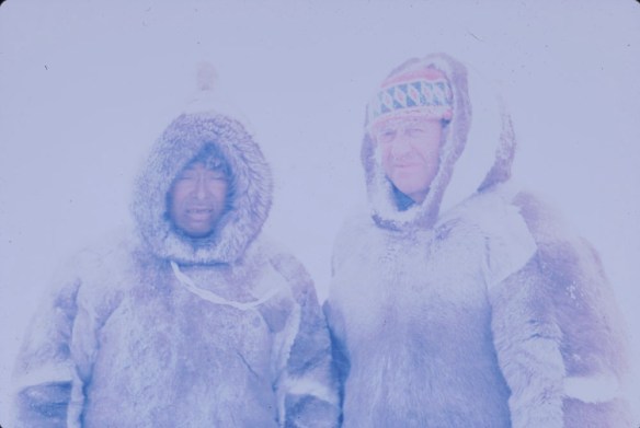Photographie couleur de Kove, un Inuit, et de Charles Gimpel, tous deux vêtus d’une parka en fourrure brun et blanc. La photo est très floue à cause d’une tempête de neige.