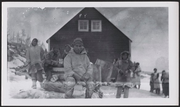 Une photographie noir et blanc d’un Innu regardant l’appareil photo, vêtu de vêtements traditionnels et assis sur une pile de provisions. À l’arrière-plan, plusieurs personnes se tiennent debout devant une maison de couleur foncée percée de deux petites fenêtres. 
