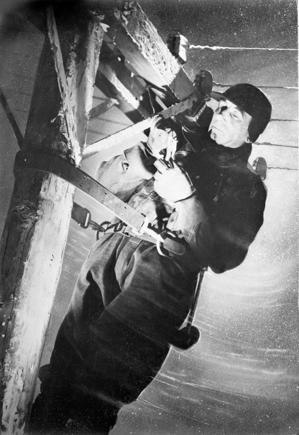 Photo noir et blanc d’un homme accroché à un poteau de téléphone, réparant des câbles sous le vent et la neige. Il porte un manteau d’hiver, des gants et un chapeau. 