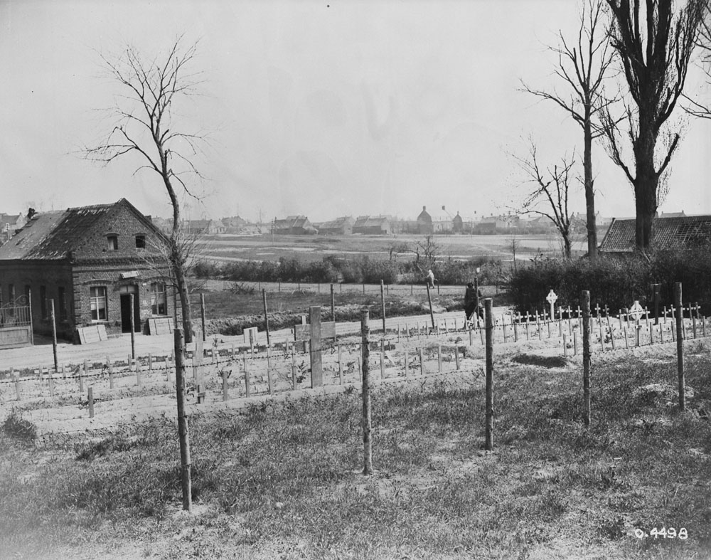 Photo noir et blanc d’un cimetière derrière une clôture, avec quelques petits arbres sans feuilles, près de Cambrai, en France. À l’arrière-plan, on aperçoit une maison et une ferme.