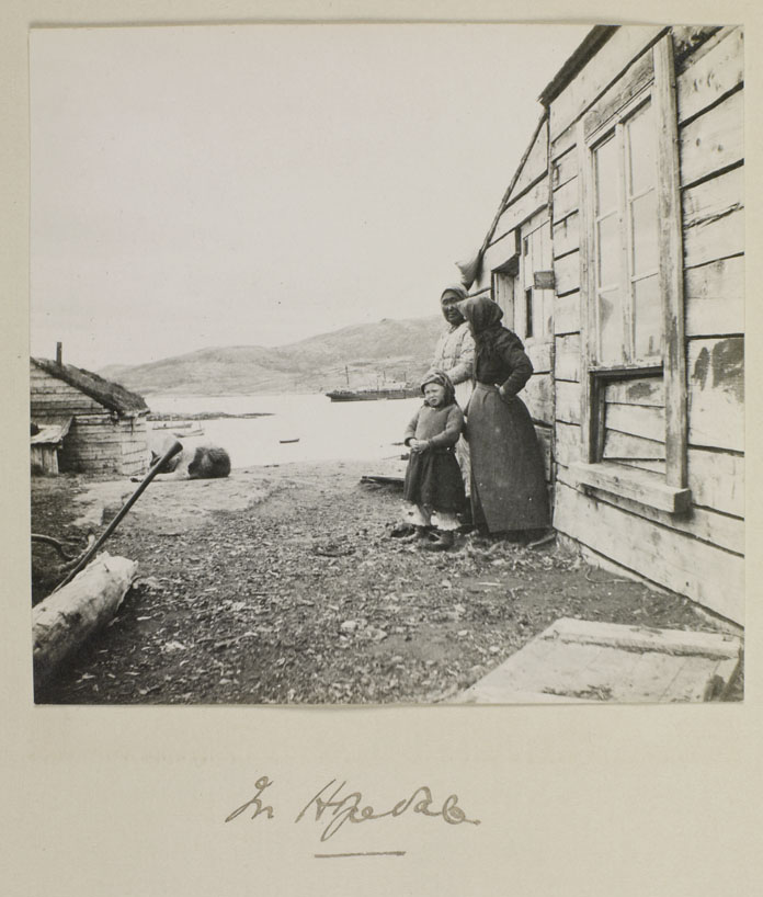 Photo noir et blanc de deux femmes et d’un enfant inuits, debout à côté d’une maison en bois.