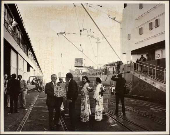 Photo montrant les dirigeants d’un port tenant des cageots de mandarines, deux femmes japonaises en costume traditionnel à côté d’une palette de cageots de mandarines ainsi que d’autres personnes et de l’équipement sur la jetée située à côté d’un navire.