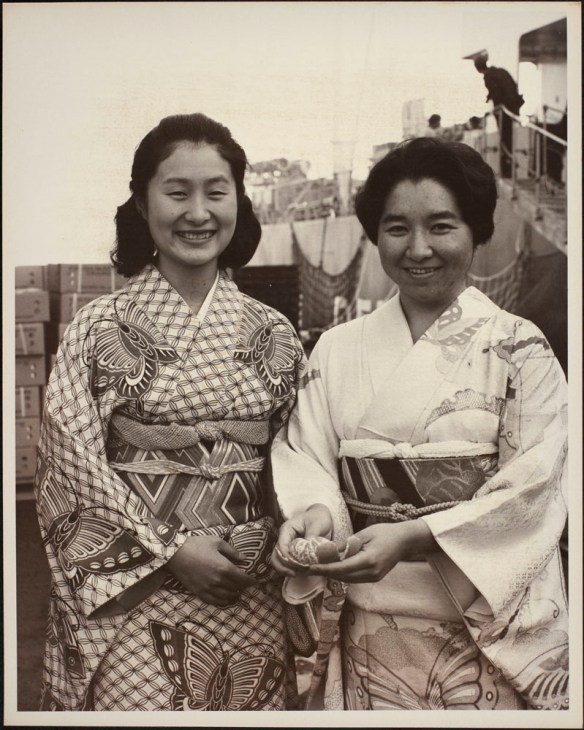 Photo de deux femmes japonaises en costume traditionnel. L’une d’elles tient une mandarine pelée. On aperçoit des cageots de mandarines et un navire à l’arrière-plan.