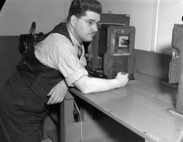 Photo noir et blanc d’un homme appuyé sur un bureau à côté d’un agrandisseur.
