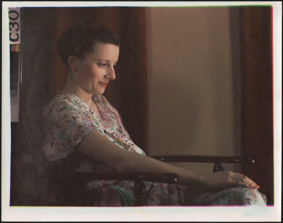 Portrait couleur d’une femme portant une robe fleurie, le visage éclairé, assise dans une chaise à appuie-bras en bois.