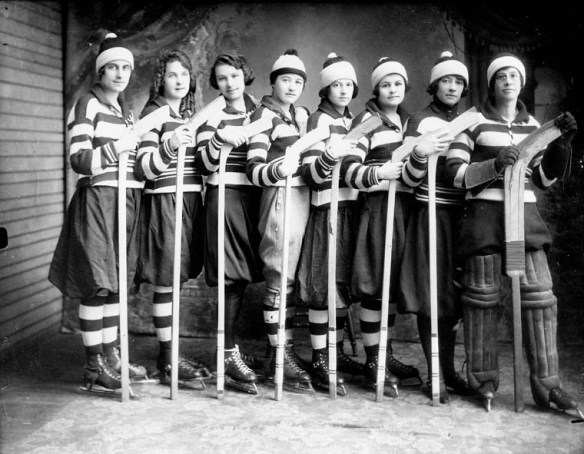 Photographie noir et blanc d’une équipe de hockey féminin. Les huit joueuses, en uniforme, sont placées côte à côte et tiennent leur bâton la palette vers le haut.