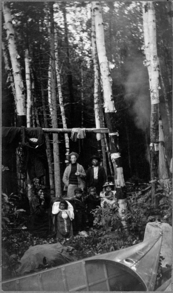 Photographie noir et blanc de huit personnes en forêt, dont un bébé dans un tikinagan. On voit un canot à l’avant-plan.