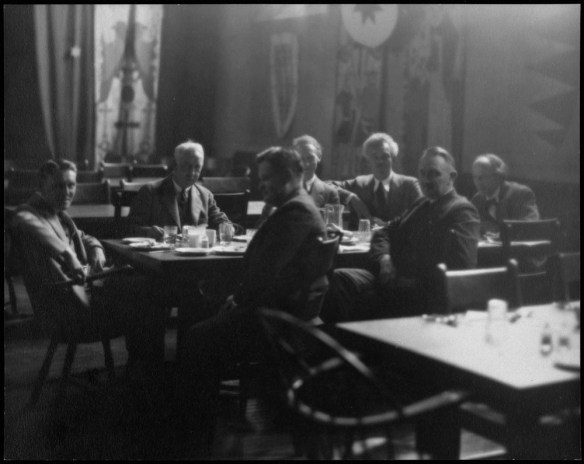 Photo noir et blanc de sept hommes vêtus de complets assis autour d’une table durant un repas. 