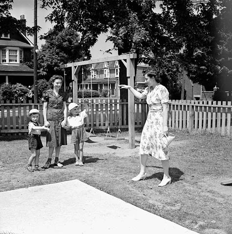 Photographie noir et blanc d’une mère saluant ses deux jeunes enfants, qui tiennent la main de leur éducatrice. La garderie semble être située sur un terrain clôturé, dans un quartier résidentiel. À l’arrière-plan figure un module avec des balançoires. 