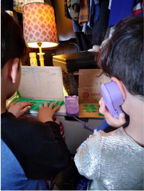 Photographie couleur de deux jeunes enfants de dos, assis à une table dans un espace encombré. Ils jouent à travailler en tapant sur des « ordinateurs » bricolés en carton et en parlant dans un téléphone en plastique. 