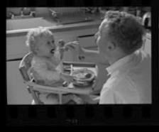 Un père fait manger son enfant, qui est assis dans une chaise haute, dans la cuisine. La bouche ouverte, il imite son enfant.