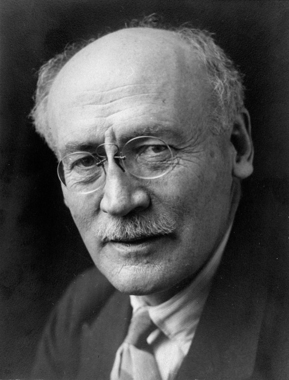 Photo noir et blanc montrant le visage d’un homme en gros plan. Il porte la moustache et de petites lunettes rondes. 