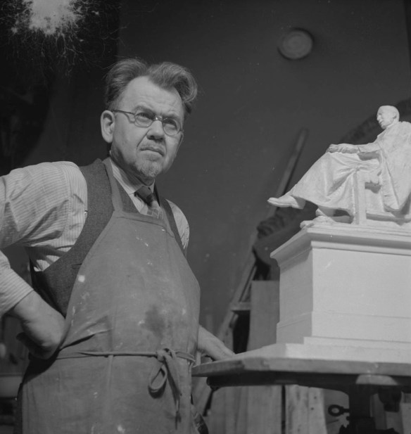 Photo noir et blanc montrant un homme portant un tablier, debout à côté d’un modèle d’une statue. Il porte des lunettes et tient un poing sur sa hanche.