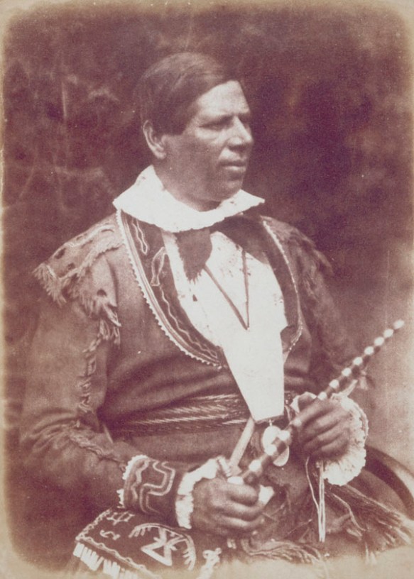 Photo sépia d’un homme assis, tenant une pipe de cérémonie et un collier de perles wampum. L’homme est vêtu d’une veste en peau de cerf; sur sa cuisse, on aperçoit un sac (également en peau de cerf) orné de motifs traditionnels. 
