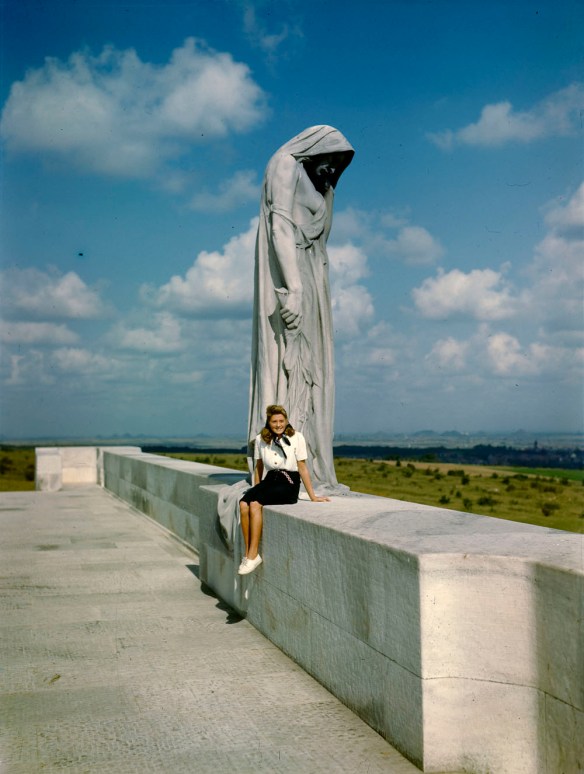 Photographie en couleur d’une jeune femme portant une blouse blanche et une jupe foncée, assise devant une grande statue représentant une femme en deuil.