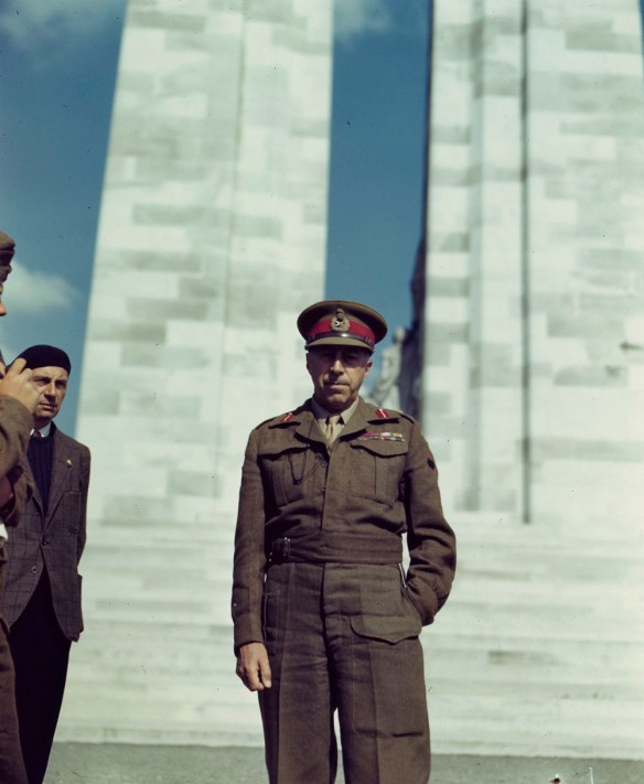 Photographie en couleurs d’un militaire en uniforme debout devant un monument commémoratif de guerre en maçonnerie. Juste à gauche, un homme portant un veston en tweed et un béret est partiellement visible.