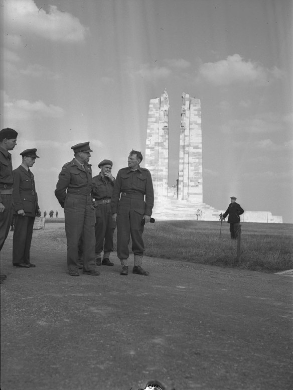 Un groupe d’hommes en uniforme militaire discutent, sous les yeux d’un civil se tenant au loin. À l’arrière-plan se dressent les deux hautes colonnes d’un mémorial de guerre en maçonnerie.