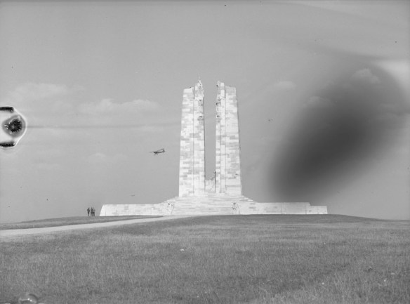 Photographie noir et blanc montrant un petit avion volant près d’un mémorial de guerre en maçonnerie caractérisé par deux hautes colonnes.