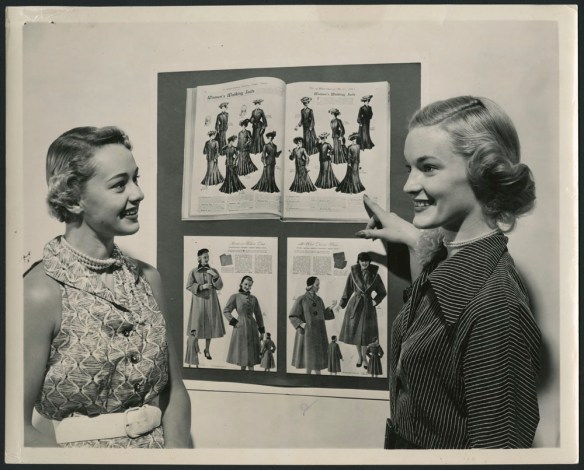 Photo noir et blanc de deux femmes, visage souriant, devant deux catalogues de mode, l’un de la fin du 19e siècle, l’autre du milieu du 20e siècle.