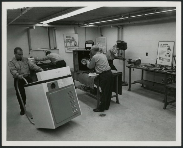 Photo noir et blanc montrant trois employés réparant des appareils dans un atelier; un quatrième employé déplace un gros appareil.