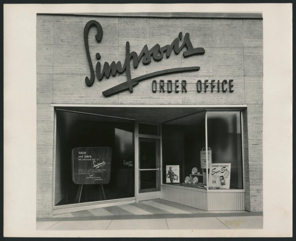 Photo noir et blanc de la façade d’un magasin de vente par catalogue. Au haut de la façade, on voit l’enseigne « Simpson’s Order Office »; des affiches publicitaires sont placées en vitrine.