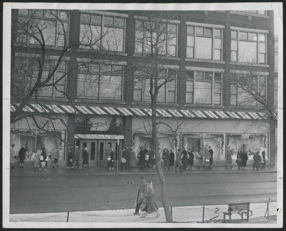 Photo noir et blanc montrant des clients devant de grandes vitrines décorées pour Noël et se dirigeant vers l’entrée principale d’un magasin Simpson’s. Des enseignes au-dessus des portes indiquent « Simpson’s » et « The Christmas Tree Store » (Boutique d’arbres de Noël).