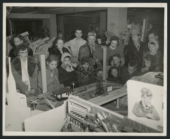 Photo noir et blanc montrant un grand groupe d’hommes, de femmes et d’enfants regardant un train électrique dans un magasin.