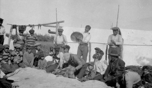 Photo noir et blanc d’une vingtaine d’hommes et de garçons, certains debout, d’autres agenouillés au sol. Au centre, un homme frappe un tambour en peau de caribou avec une baguette de bois. Il porte une chemise blanche et un pantalon sombre, et tient une pipe en bois à la bouche. À l’arrière-plan, on voit une tente rectangulaire en toile blanche, et de la viande qui sèche sur un poteau de bois.