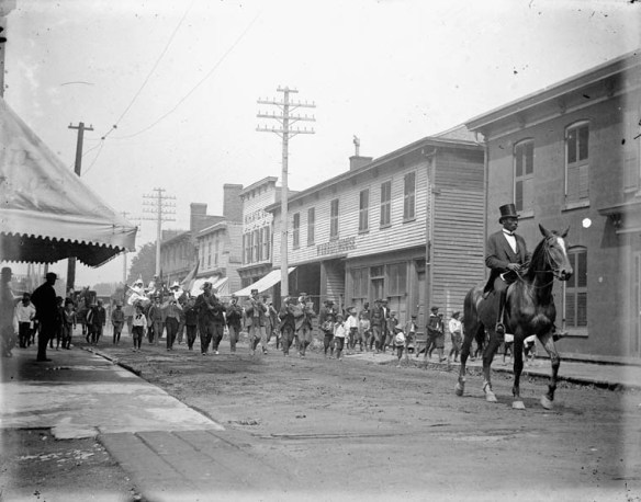 Une foule défile au milieu d’une rue dans une petite ville. Des magasins en bois bordent la rue de chaque côté. La foule est menée par un homme noir distingué, moustachu, vêtu d’un haut-de-forme et d’une queue-de-pie, qui se déplace à cheval. Une fanfare, des groupes de petits garçons et plusieurs adultes marchent derrière lui. La plupart des gens dont le visage est visible dans la foule semblent Noirs. En arrière-plan, un deuxième cheval tire un char sur lequel se tiennent des femmes vêtues de robes blanches et de chapeaux à large bord.