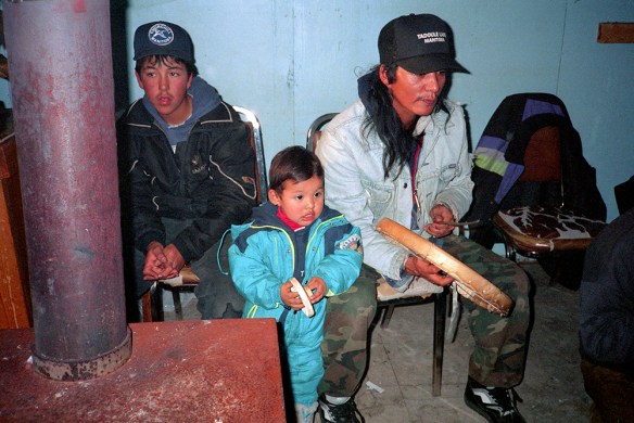 Photo couleur d’un homme, d’un adolescent et d’un bambin regardant une partie de jeux de mains dénés. L’homme frappe un tambour en peau de caribou avec une baguette de bois. Le bambin tient son propre petit tambour. 