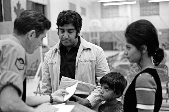 Photographie noir et blanc d’un homme en uniforme qui regarde un papier, devant un homme tenant des documents et vêtu d’une chemise foncée sous une veste claire, un jeune garçon, et une femme aux cheveux attachés en queue de cheval.