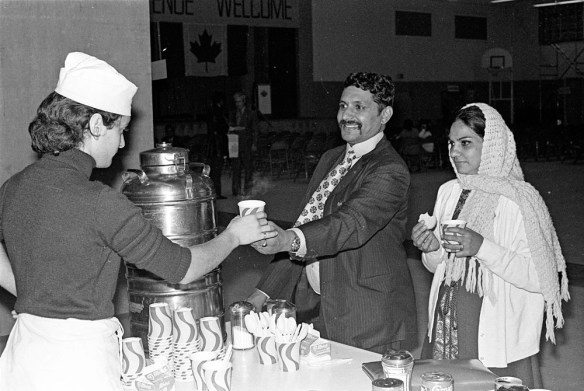 Photographie noir et blanc d’une femme portant un tablier et un calot de cuisine qui tend un verre en carton à un homme souriant en complet-veston, accompagné d’une femme portant un foulard et tenant un biscuit ainsi qu’un verre de carton.