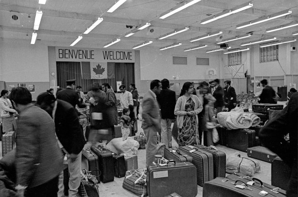 Photo noir et blanc d’une grande salle relativement bondée. Des valises et bagages divers jonchent le sol. Sur le mur, un drapeau canadien est surmonté d’une banderole disant « Bienvenue, Welcome ».