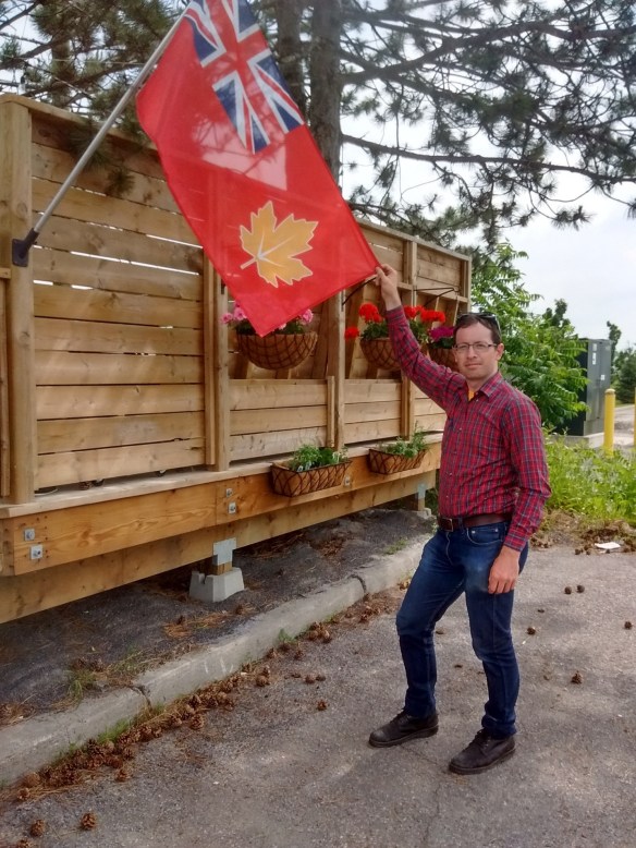 À l’extérieur, un homme en jeans et en chemise à carreaux rouge fait face à l’objectif, debout près d’un drapeau qu’il tient par le coin.