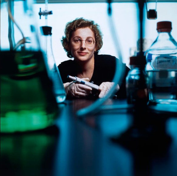 Photo couleur d’une femme avec des lunettes, face à l’objectif. On aperçoit des récipients et des bouteilles remplis de liquide à l’avant-plan. 