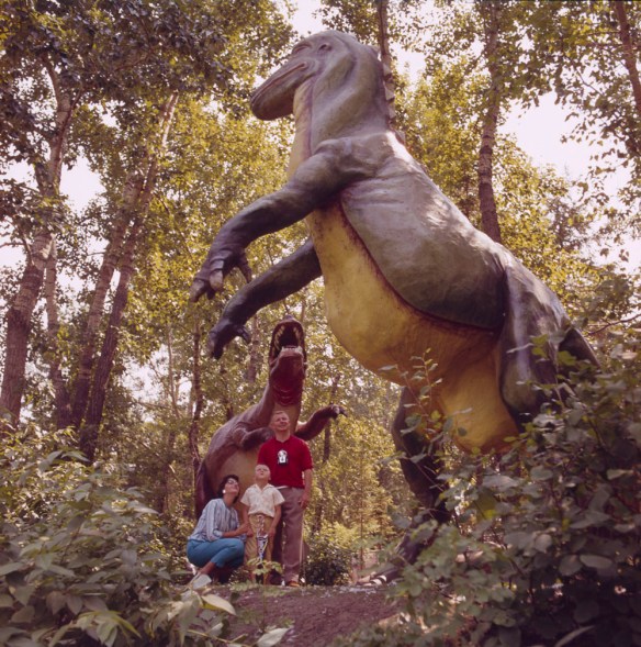 Une femme, un enfant et un homme sous l’imposante sculpture d’un dinosaure entourée d’arbres. Le groupe regarde un autre dinosaure. 