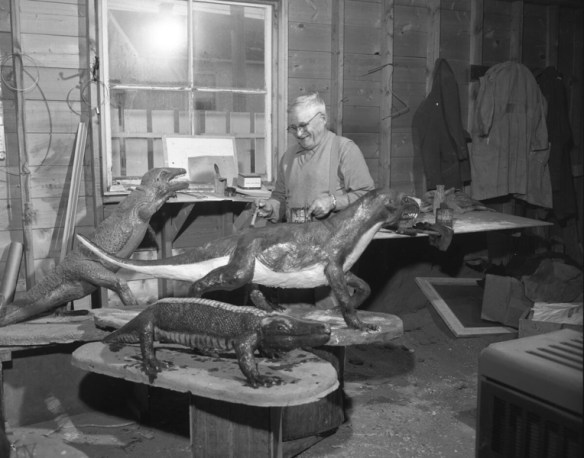 Un homme dans un garage ou un atelier, près de trois sculptures de reptiles préhistoriques. Il tient dans ses mains une canne de peinture et un pinceau. 