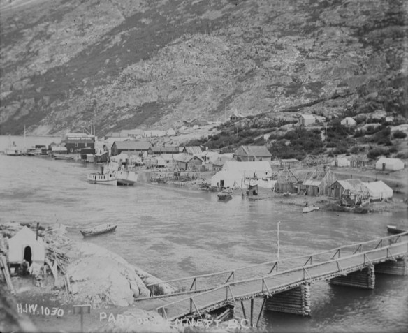 Photographie en noir et blanc de la ville de Bennett, en Colombie-Britannique, au bord du lac Bennett. Il y a des bâtiments et des structures temporaires le long du rivage, avec un flanc de montagne en arrière-plan et un pont en bois au premier plan.