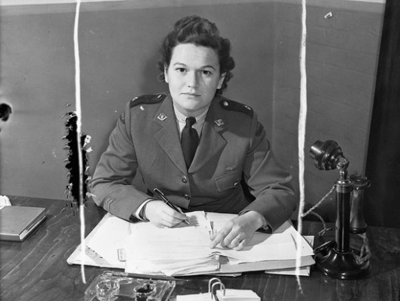 Photo noir et blanc d’une femme en uniforme militaire regardant l’objectif. Elle est assise derrière un bureau et tient un stylo dans sa main droite. Un téléphone de type chandelier est posé sur le bureau, à sa gauche.