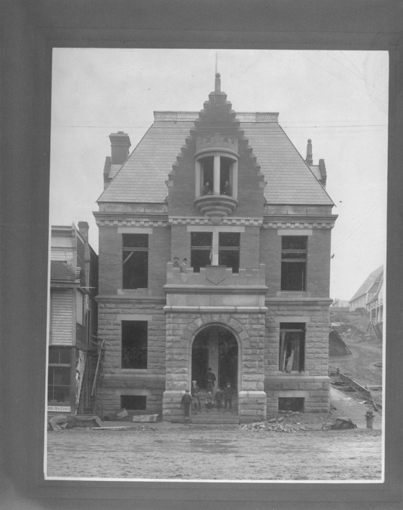 Photographie en noir et blanc de l’extérieur du bureau de poste. De petits groupes d’hommes et d’enfants se tiennent devant l’entrée inachevée du bâtiment et l’une des fenêtres toujours en construction.