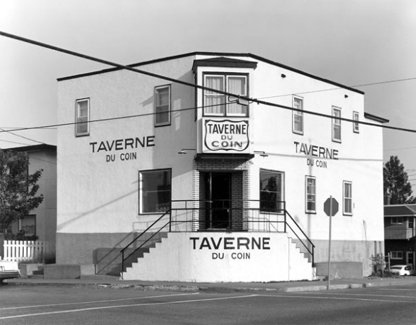 Photo noir et blanc d’un bâtiment de couleur blanche, situé au coin d’une rue et flanqué de deux escaliers menant à la porte d’entrée. Sur le bâtiment, on peut lire à quatre endroits l’inscription « Taverne du coin ». Un panneau d’arrêt se trouve devant le bâtiment. 