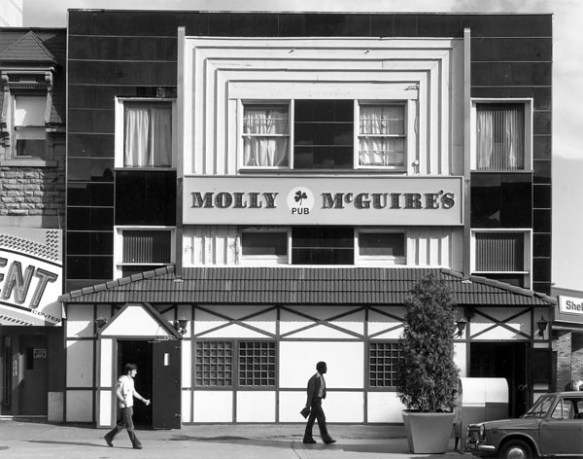 Photo noir et blanc d’un bâtiment à la façade blanche et foncée. Les mots « Molly McGuire’s Pub » (avec un petit trèfle) y sont inscrits. À l’avant-plan, on voit deux hommes et une voiture. 