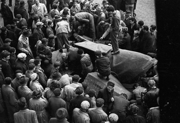 Photo noir et blanc d’une foule entourant un monument renversé. Des hommes se tiennent debout sur le monument, regardant vers le bas. 