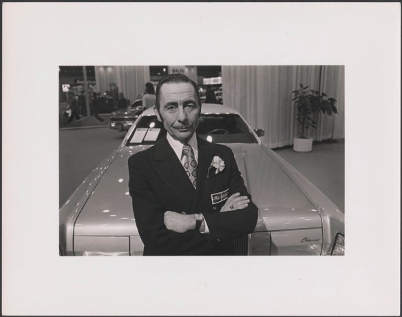 Photo noir et blanc d’un homme en complet, portant un œillet blanc au revers de sa poche et s’appuyant sur le capot d’un modèle de voiture des années 1970. L’homme a les bras croisés et sourit à l’objectif. 