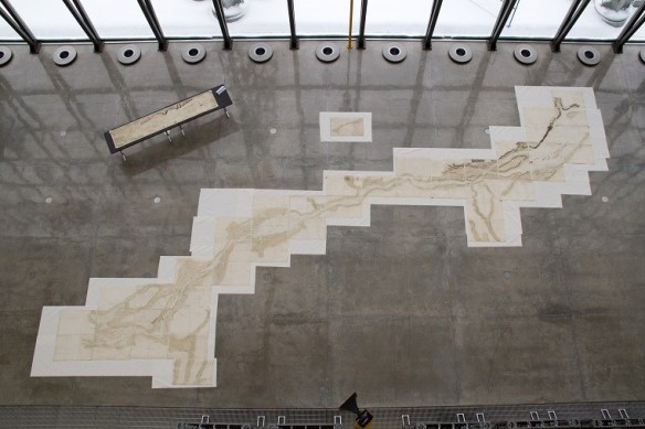 Photo couleur d’une grande carte manuscrite, composée d’une quarantaine de feuilles, étalée sur un carrelage sombre. Une carte rectangulaire plus petite se trouve sur une table dans le coin supérieur gauche.