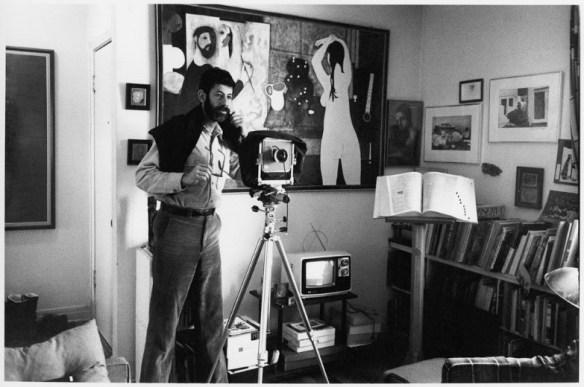Photo noir et blanc d’un homme portant une chemise boutonnée, un chandail jeté sur ses épaules. L’homme se tient debout derrière un appareil photo reposant sur un trépied. 