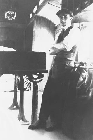 Photographie en noir et blanc d’un jeune homme à côté de tables et de sacs postaux dans une voiture postale.